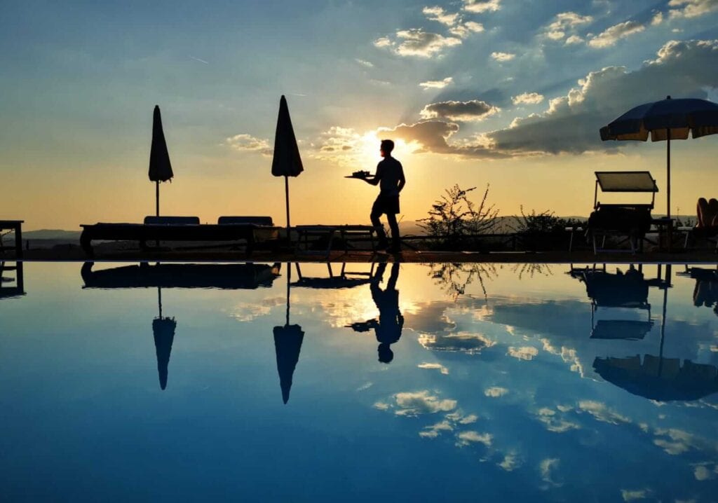 Silhouette di una persona vicino a una piscina al tramonto, con ombrelloni e lettini riflessi nell'acqua.